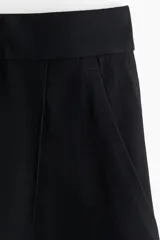 Pantalón sastrero negro de sarga con pinzas, corte holgado, tiro alto, pretina prolongada con botón, bolsillos al bies y bolsillos insertados atrás. Piernas anchas con pliegues adelante y atrás.