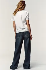 Camiseta blanca de algodón con cuello redondo y mangas cortas. Estampado frontal con texto "Meet me at Palais Lomtam, 7, Passage des Rêveries" enmarcado en un diseño ondulado.