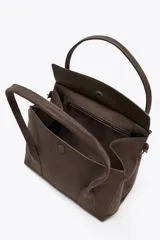 Bolso marrón de gamuza sintética, tipo city bag, con doble asa de hombro y cierre con solapa e imán. Tiene correa ajustable con hebillas metálicas en los laterales y bolsillo interior con cremallera.
