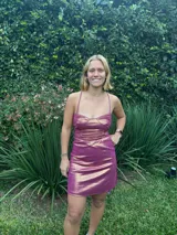 Vestido corto color fucsia con textura brillante, breteles finos y escote recto. Presenta corte tipo corsé con costuras fruncidas y falda ajustada.