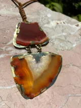 Collar con colgante de dos piedras naturales irregulares engarzadas en metal plateado y dorado, suspendidas de un cordón de cuero marrón con detalle de nudo corredizo. Las piedras tienen vetas en tonos naranja, marrón y blanco.