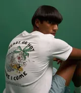 Remera blanca de algodón con cuello redondo y manga corta. Estampa en la espalda con diseño de esqueleto surfeando una ola junto a una palmera.