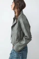 Campera biker color verde grisáceo con efecto desgastado, cuello con solapa y cierre lateral. Presenta herrajes plateados y detalles de cordones entrelazados en los codos y en la espalda.