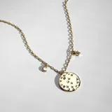 Collar de cadena fina dorada con dije principal circular con relieve y cristales incrustados, acompañado de dijes colgantes de luna creciente y estrella, también con cristales.