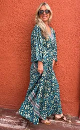 Vestido largo estilo cruzado (wrap dress) de manga larga acampanada, con estampado floral en tonos verde petróleo, amarillo pálido y celeste. Presenta un ribete decorativo en el escote y el borde del cruce.