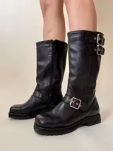 Bota de caña media de cuero vacuno color marrón oscuro, con punta cuadrada, cierre lateral y suela track negra. Presenta detalles de hebillas metálicas plateadas en la caña y en la parte inferior del empeine.