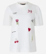 Remera blanca de algodón con cuello redondo y mangas cortas abullonadas. Presenta estampados de un lazo, una flor, un corazón y frases caligráficas.
