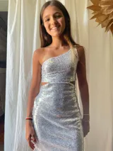 Vestido corto asimétrico con un hombro descubierto y dos breteles finitos que se atan cruzados en la espalda. El vestido es de lentejuelas plateadas y tiene un corte en la cintura.