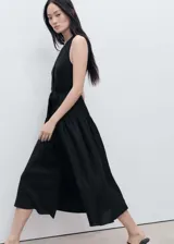 Vestido midi negro sin mangas, confeccionado en lino. Presenta escote en V, cierre frontal con botones y cinturón de lazo. La falda es acampanada con un volante en el ruedo.