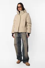 Campera puffer beige de corte oversize, confeccionada en textil impermeable. Cuenta con capucha forrada con tancas y elástico, cuello alto, cierre frontal con cartera y abrojo, bolsillos ribete con cierre, mangas desmontables con mitones y cintura ajustable con elástico y tancas. Incluye cinta gross estampada para transportarla al hombro y bolsillo interno con cierre invisible.