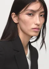 Blazer negro de diseño entallado, con solapas de muesca, hombros estructurados y cierre frontal de un solo botón. Presenta dos bolsillos de ribete delanteros y un bolsillo de ribete en el pecho.