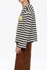 Camiseta de tejido de punto de manga larga, con patrón de rayas horizontales anchas en blanco crudo y negro. Presenta cuello redondo con ribete negro y un pequeño bordado amarillo en el pecho.