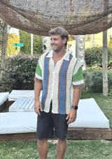 Camisa de manga corta tejida a crochet, con cuello polo y botones frontales. Presenta un diseño de rayas verticales en blanco, azul, verde y marrón.