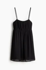 Vestido corto negro de tejido transparente con forro. Tiene breteles spaghetti, escote cuadrado con moña decorativa de satén adelante y falda evasé. El cuerpo y la cintura tienen fruncido elástico.