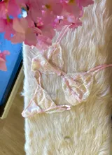 Conjunto de lencería de encaje floral en tonos rosa y crema, compuesto por un corpiño triangular sin aro con breteles finos regulables y una tanga regulable.