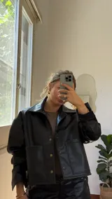 Campera corta negra de ecocuero con cuello camisero, cierre frontal con botones a presión, dos bolsillos con solapa y mangas largas con puños abotonados.