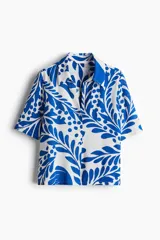 Camisa de manga corta con estampado de hojas y flores en tonos azules sobre fondo blanco. Tiene cuello clásico, botones frontales, corte holgado y canesú con pliegue en la espalda.