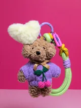 Llavero con forma de oso de peluche marrón con sweater tejido color lila, detalle de cerezas bordadas y corazón de peluche blanco.