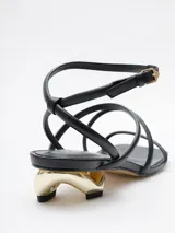 Sandalias negras de cuero con tiras finas, taco geométrico dorado y pulsera al tobillo.