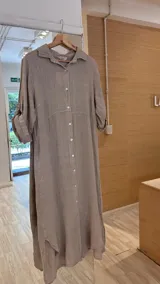 Vestido camisero largo de lino color beige/taupe, con cuello camisero, manga larga enrollada y cierre frontal de botones de nácar. Presenta un corte en la cintura y bajo asimétrico con aberturas laterales.