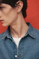Camisa de denim azul de algodón rígido, corte holgado, cuello inglés, tapeta clásica con botones blancos, bolsillo de parche en el pecho, canesú en la espalda, mangas largas con puños abotonados y bajo levemente redondeado.