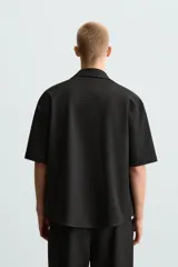Sobrecamisa negra de tejido con elasticidad, cuello solapa y manga corta. Tiene dos bolsillos de plastrón en el pecho y cierre frontal con botones a presión.