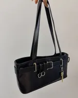 Cartera de hombro negra, de forma rectangular y acabado liso. Presenta detalles de hebillas plateadas y tiras decorativas en el frente, junto con un dije colgante en forma de llave dorada.