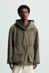 Parka color verde militar de corte amplio, confeccionada en tejido técnico ligero. Cuenta con cuello alto con capucha ajustable, cierre frontal con botones, mangas largas con puños ajustables, bolsillos de plastrón con solapa en la parte delantera, cintura ajustable con cordón y bajo con aberturas laterales ajustables con cordón.