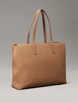 Cartera tote Calvin Klein color tostado, de tamaño mediano, confeccionada en cuero sintético granulado con textura de logo monograma en el bolsillo delantero. Tiene asas fijas, cierre magnético, bolsillo exterior, compartimento interior con cremallera y colgante extraíble con logo.