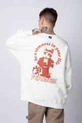 Buzo color crema de corte oversize confeccionado en tela friza con cuello redondo y puños y cintura acanalados. Presenta un pequeño bordado en el frente y un estampado grande en la espalda.