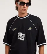 Remera deportiva de algodón, color negro, con corte oversized, cuello redondo y mangas cortas. Presenta ribetes contrastantes en blanco en el cuello y los bordes de las mangas. Tiene un estampado localizado en el pecho con tipografía en relieve y un pequeño logo en el lateral superior.