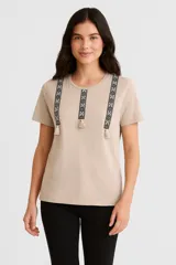 Remera color beige con cuello redondo y mangas cortas. Presenta apliques bordados con motivos geométricos y borlas en el frente.