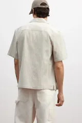Camisa de manga corta color beige claro, confeccionada en tejido liviano de lino y algodón. Tiene cuello resort, tapeta lisa, bolsillo superior y bajo recto con aberturas laterales.