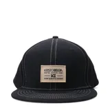 Gorro estilo snapback negro con visera plana y costuras blancas contrastantes. Parche frontal rectangular color beige con texto negro que dice "KRISTOBELGA ESTD. SINCE 1981 - KB - SURF & SKATE GOODS".