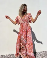 Vestido largo de corte evasé con estampado floral en tonos rosados y anaranjados. Tiene escote en V cruzado, mangas cortas con volantes y falda con volantes asimétricos.