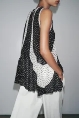 Top de cuello redondo y manga sisa con detalle de tejido plisado semitransparente y estampado de lunares blancos y negros. Bajo asimétrico y cierre en espalda con abertura y botón.