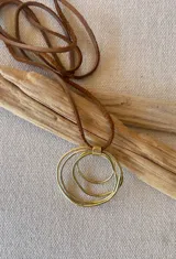 Colgante con dije de tres círculos concéntricos de metal dorado, unidos por una pieza rectangular. Cordón fino de cuero marrón.