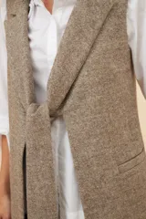 Chaleco largo color marrón claro, abierto, con lazo en la delantera y bolsillos ojal. Confeccionado en lana fina sin teñir.