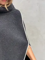Poncho tejido de punto grueso color gris con cuello alto y aberturas laterales con ribete blanco.