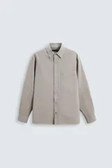 Camisa color gris topo de corte relaxed fit, confeccionada en tejido de mezcla de algodón suave. Tiene cuello solapa, manga larga con puños abotonados, bolsillo de parche en el pecho y cierre frontal con botones.
