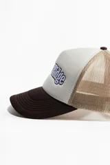 Gorra tipo trucker con visera curva color marrón, frente blanco con bordado de la marca "Raptor Ridge" en azul y blanco, y rejilla color beige.