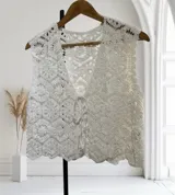 Chaleco sin mangas de crochet blanco, con tejido calado de patrón geométrico y borde festoneado. Presenta un escote en V profundo que se anuda al frente con un lazo fino.