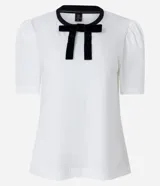 Blusa blanca de media malla de algodón, con cuello redondo con ribete negro y moña de terciopelo negra. Tiene mangas cortas abullonadas.