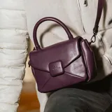 Cartera bandolera color púrpura de cuero sintético acolchado, con asa corta fija y correa larga regulable. Tiene acceso con solapa, un bolsillo interno con cierre, un bolsillo externo abierto y herrajes plateados.