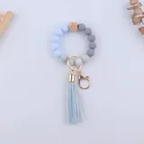 Llavero pulsera con cuentas de silicona en tonos celeste, gris y blanco, con un cubo de madera y flecos celestes.