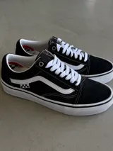 Championes Vans Skate Old Skool, color negro con franja lateral blanca, cordones blancos y suela blanca con línea negra. Presentan un detalle de bandera a cuadros en el lateral.