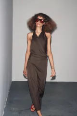 Vestido largo color marrón oscuro, de cuello halter y escote fluido. Tiene hombros descubiertos, detalle de pliegues laterales, forro interior a tono y cierre lateral con cremallera oculta y en espalda con botones.