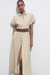 Vestido midi camisero color beige, confeccionado en lino. Presenta cuello solapa, mangas cortas con vuelta, jaretas delanteras, bolsillos laterales ocultos en costura, cierre frontal con botones y cinturón trenzado marrón.