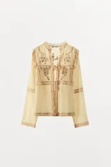 Blusa color crema de algodón y viscosa, con cuello mao y mangas largas. Presenta bordados florales con abalorios y terminaciones en puntilla. Se cierra con lazos en el frente.