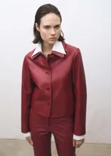 Chaqueta de cuero ovino color borgoña, de corte recto, con cuello camisero blanco, cierre frontal con botones y dos bolsillos de parche con solapa.